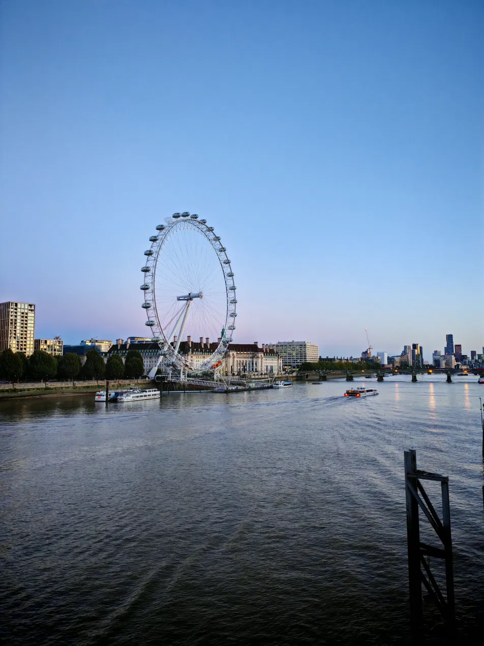 The London Eye and County Hall at dusk seen from across the Thames, with river boats and the South Bank skyline behind