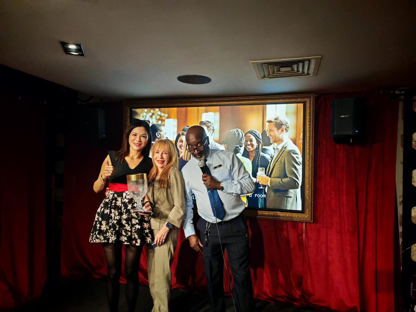 Gina Cheng holding the crystal goblet beside a winner of the business-card draw and Lee from the Grosvenor Rialto team, on stage at the V8 Nexus October Members' Evening