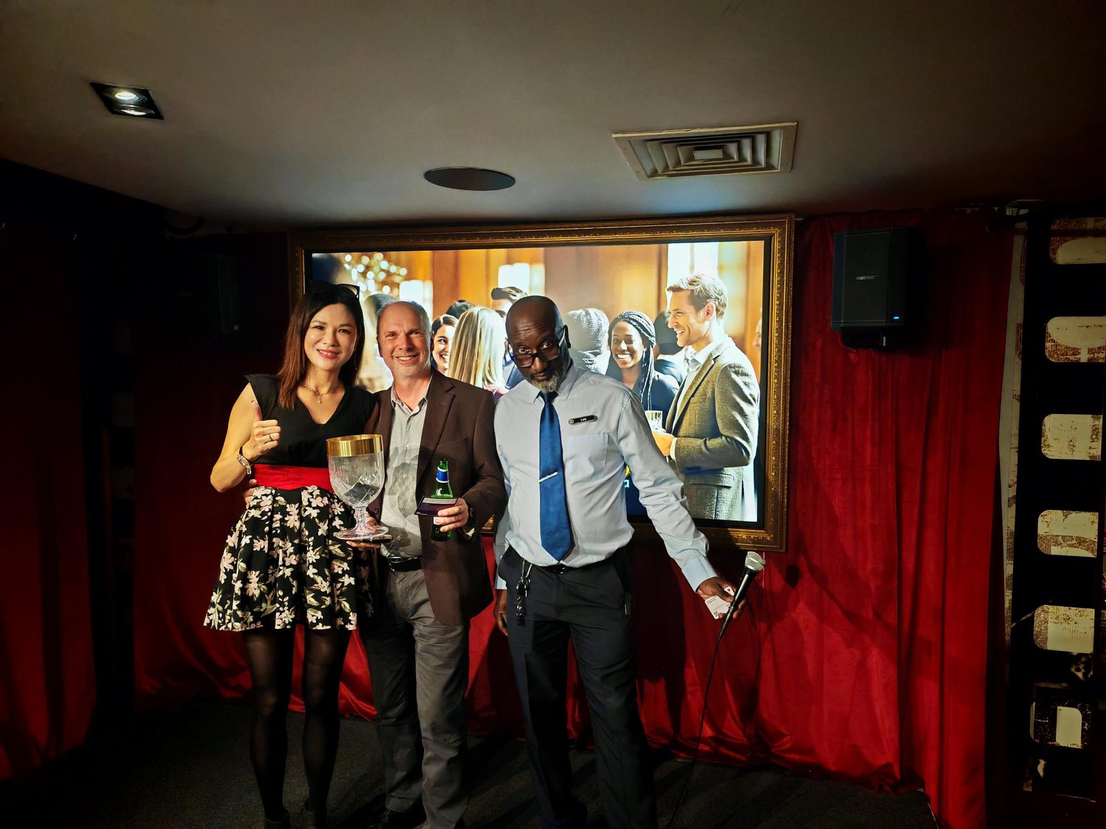 Gina Cheng opening the V8 Nexus October Members' Evening on stage in front of a London skyline slide reading V8 Nexus, London Executive Circle, at the Backstage Bar, Grosvenor Rialto, Piccadilly
