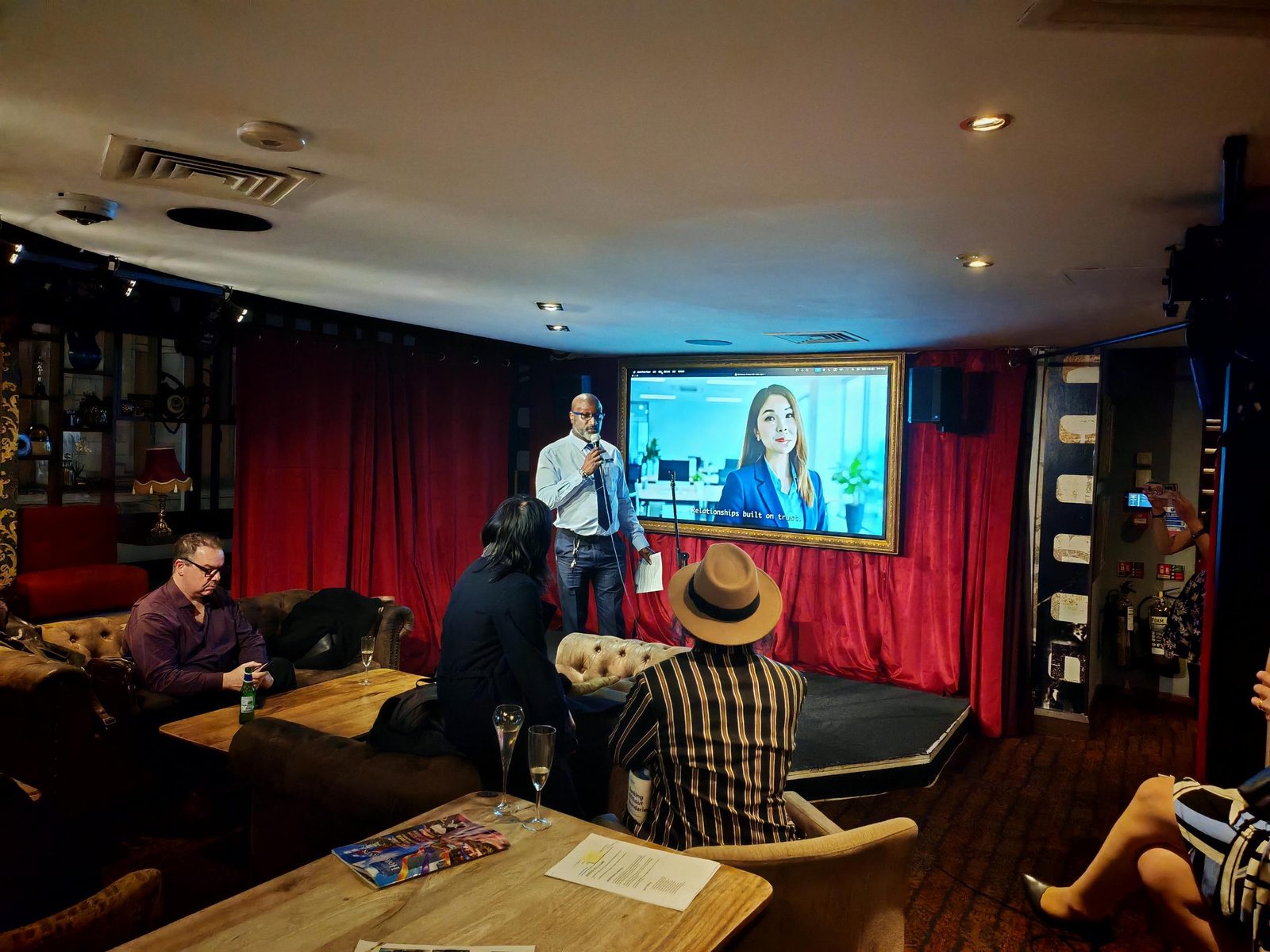 Guests at the V8 Nexus October Members' Evening watching the members' film on the gold-framed screen at the Backstage Bar, Grosvenor Rialto, with the caption Relationships built on trust